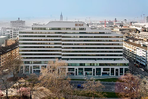 Eines der sanierten Gebäude von HGP Architekten als Architekturbüro Fulda. Das Hochhaus ist eher breit als hoch, weiß und hat viele Reihen an Fenster, die rund um das Gebäude verlaufen. Im Hintergrund sind weitere Gebäude, Straßen und Bäume zu sehen.